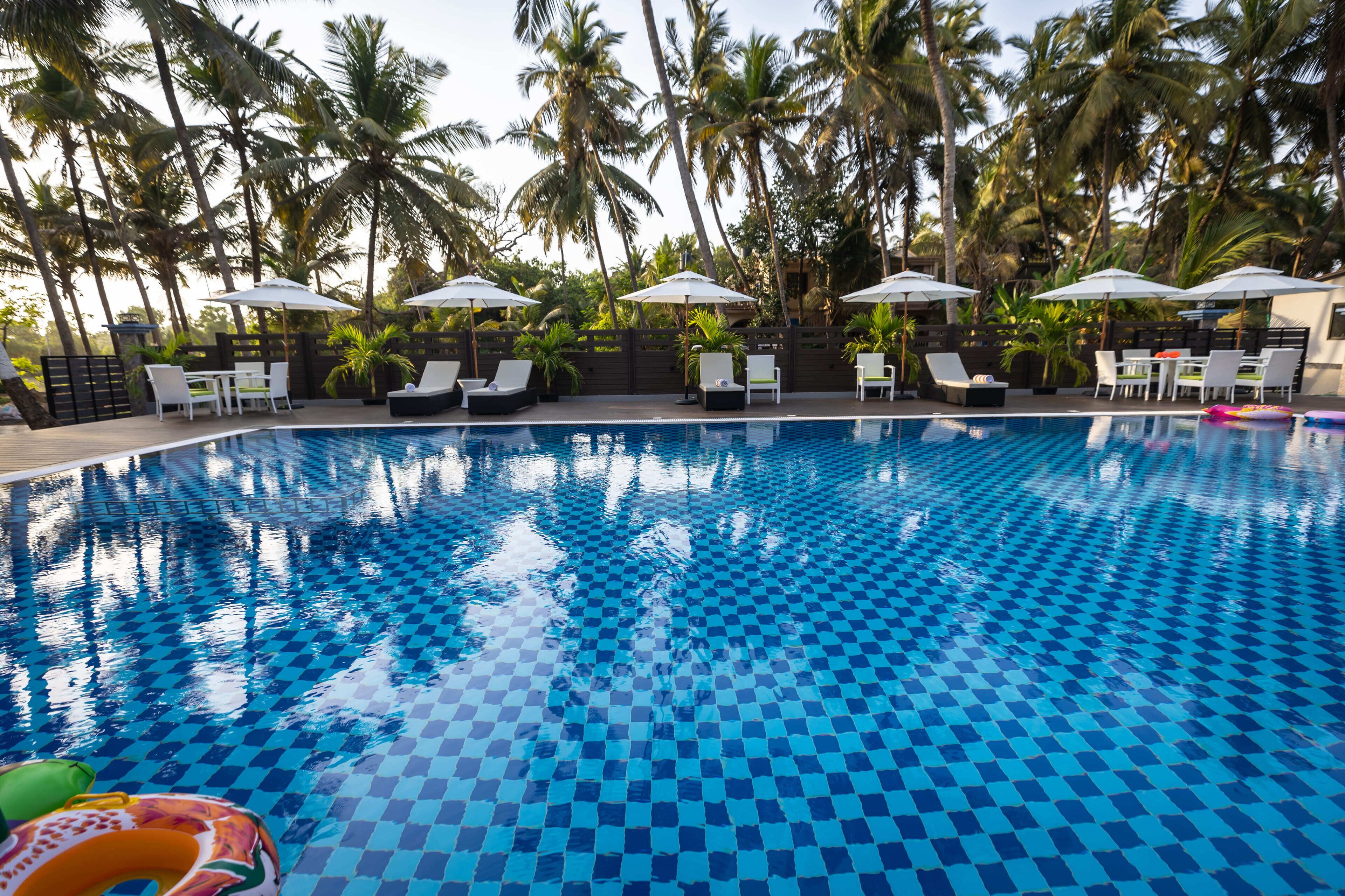 Una piscina con baldosas a cuadros, rodeada de sillas de descanso y sombrillas, con palmeras al fondo.