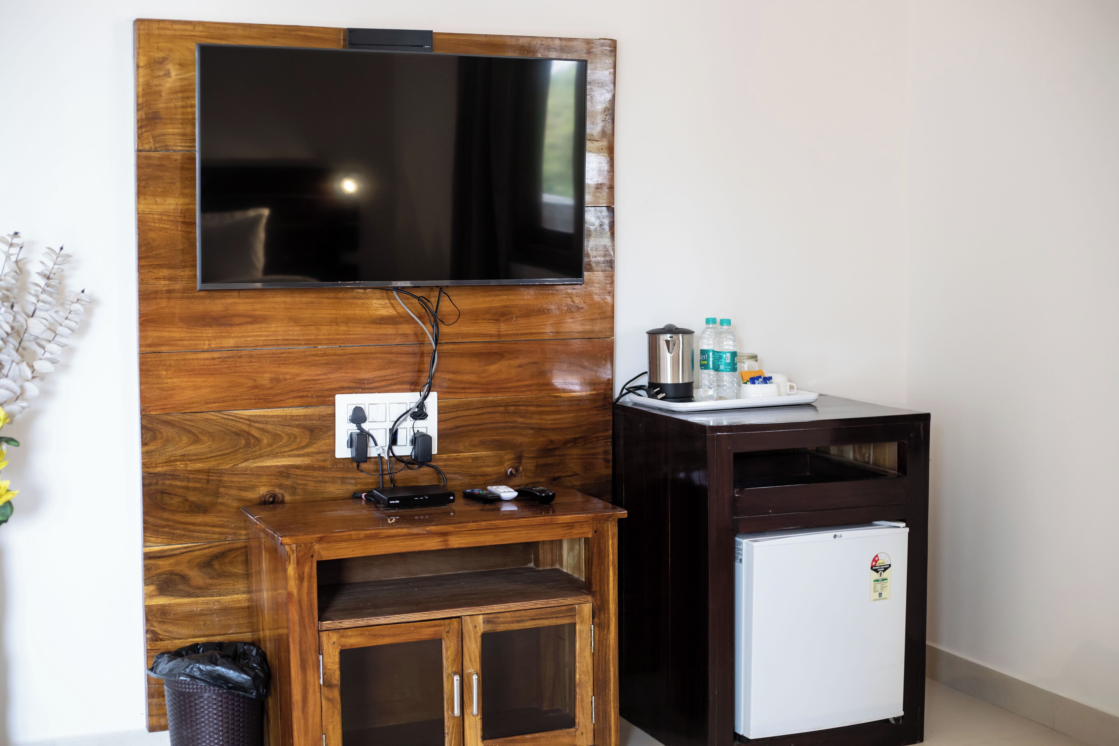 Une pièce avec un téléviseur fixé au mur, un petit meuble en bois contenant une poubelle, un réfrigérateur blanc et une petite table avec une bouilloire et des bouteilles.