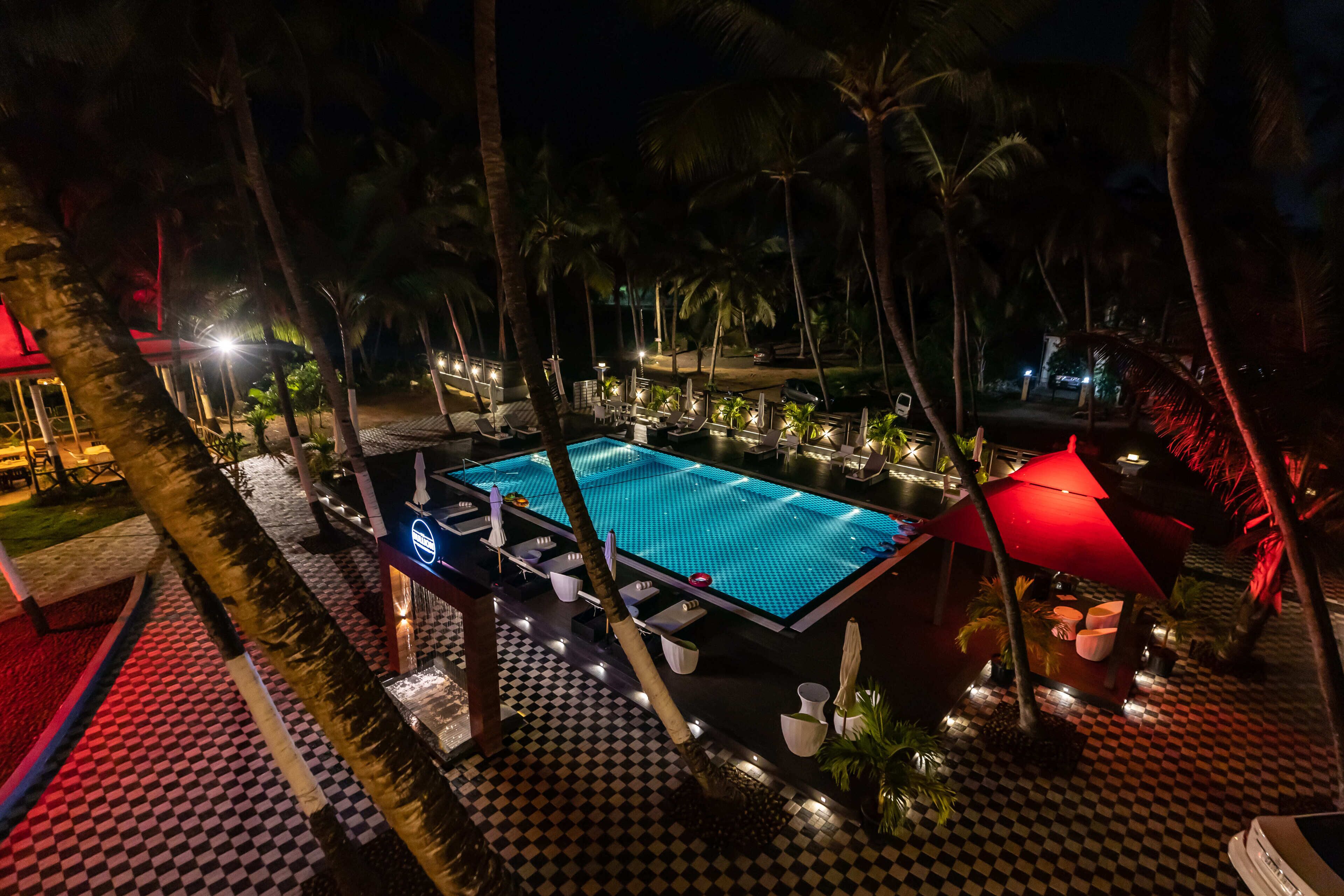 A nighttime view of a pool area with lounge chairs and umbrellas, surrounded by palm trees and a checkered floor pattern.