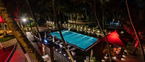 A nighttime view of a pool area with lounge chairs and umbrellas, surrounded by palm trees and a checkered floor pattern.
