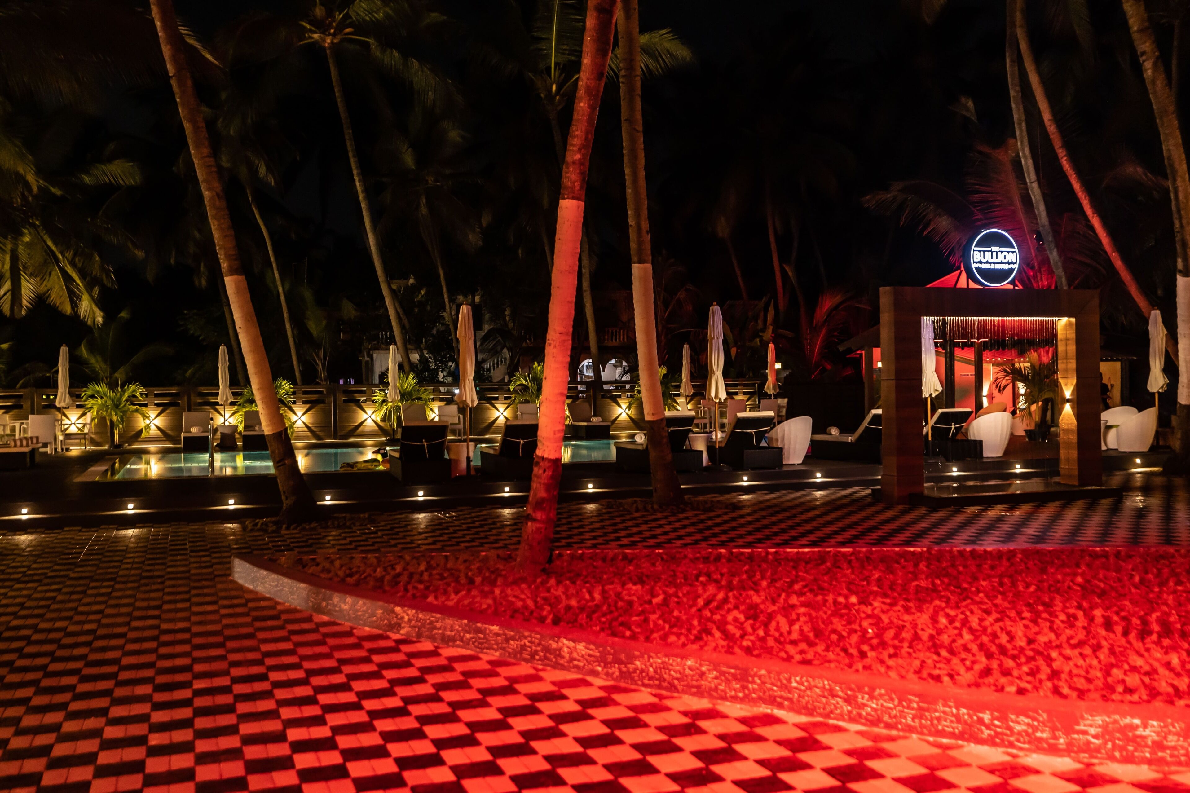 Vue nocturne d’une piscine d’hôtel, avec un sol à motifs en damier, des palmiers et un panneau lumineux au néon indiquant le mot « BULLION ».