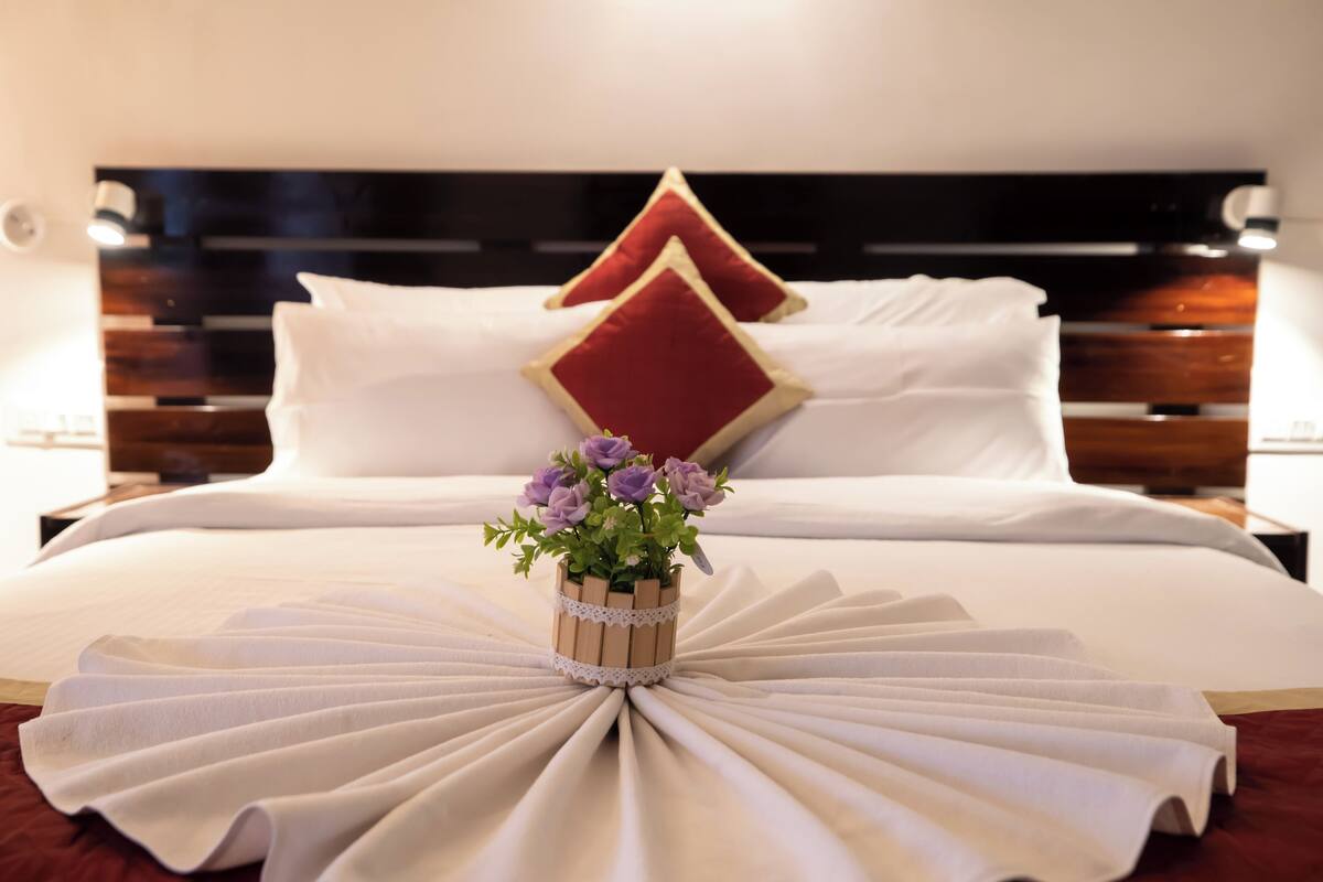 Una cama bien hecha con sábanas blancas, dos almohadas rojas y un pequeño jarrón de madera con flores moradas.