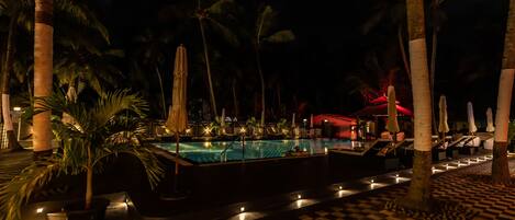 Zona de piscina nocturna con suelo a cuadros, palmeras y sillones de descanso.