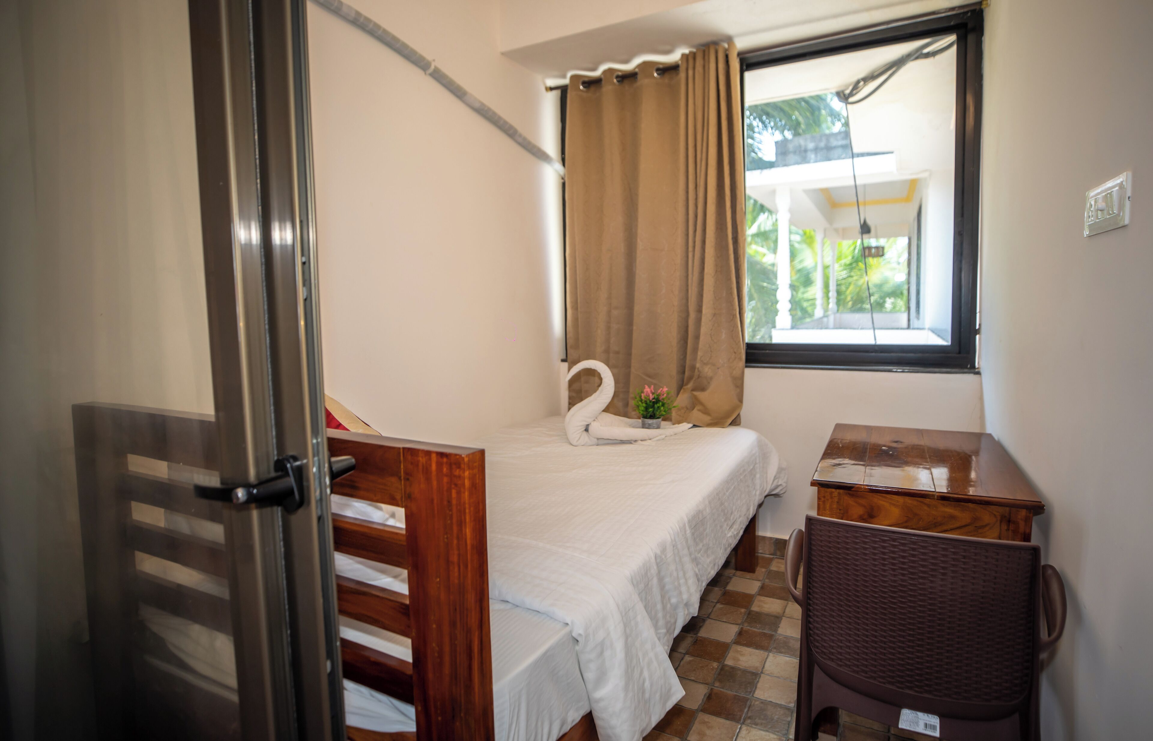 Una habitación pequeña y limpia con cama, una silla, una mesita y una ventana con cortinas.