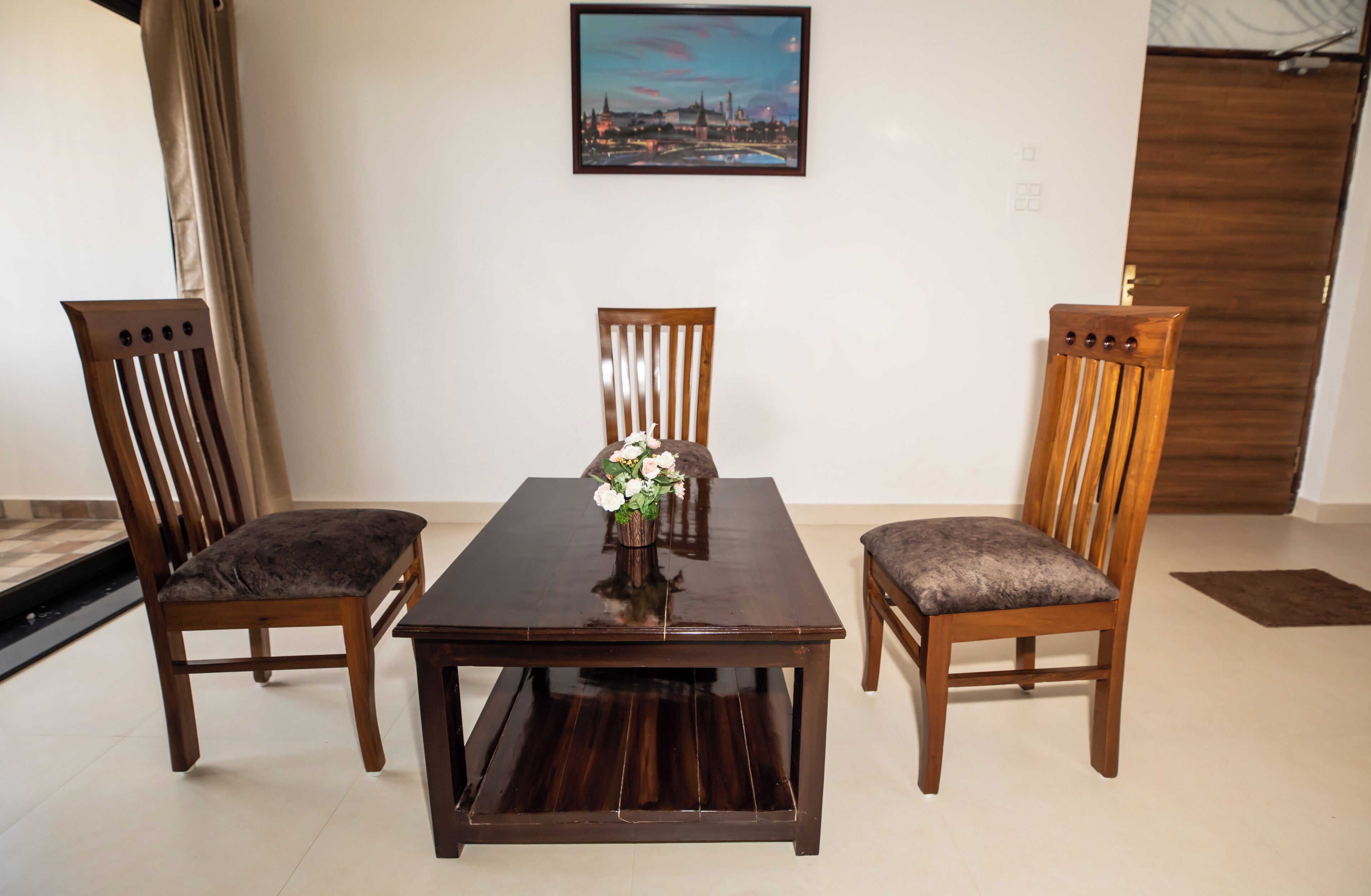 A dining area with a wooden table and chairs, a vase with flowers, and a cityscape painting on the wall.