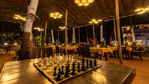 An outdoor dining area with tables set for a chess game, surrounded by chairs and a thatched roof structure. - The Woodside Inn Goa (Benaulim)