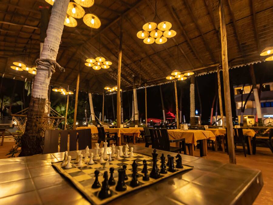 An outdoor dining area with tables set for a chess game, surrounded by chairs and a thatched roof structure.