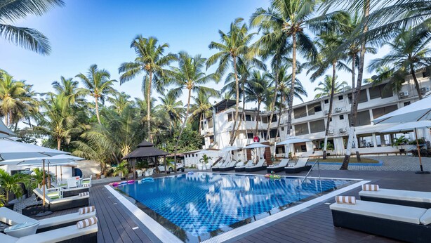 A pool area with lounge chairs and umbrellas, surrounded by palm trees and a building with multiple balconies. - The Woodside Inn Goa (Benaulim)