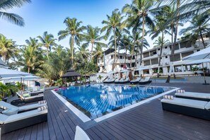 A pool area with lounge chairs and umbrellas, surrounded by palm trees and a building with multiple balconies. - The Woodside Inn Goa (Benaulim)