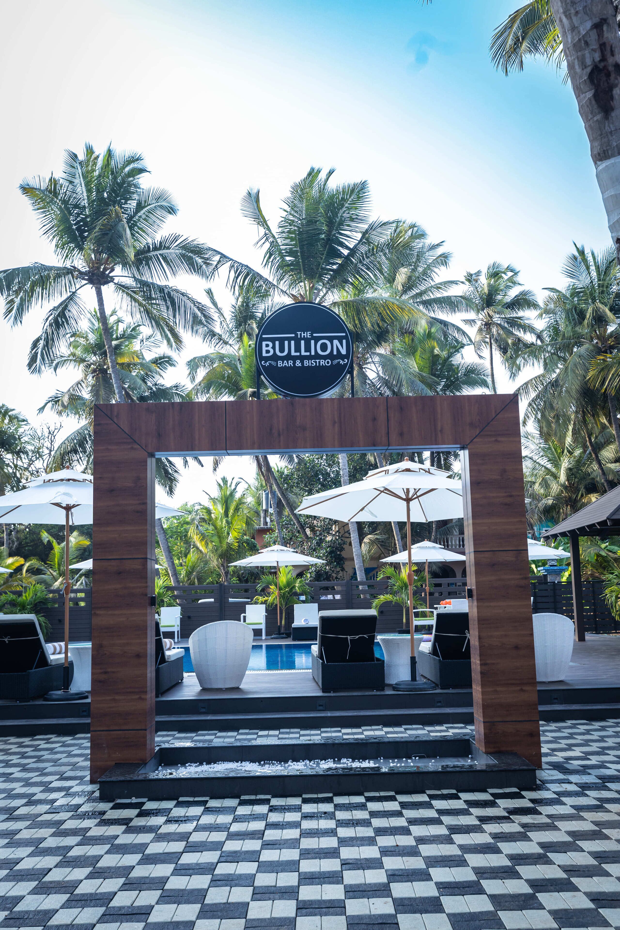 Un arco de madera con un letrero que dice "The Bullion Bar & Bistro" frente a un área de piscina con sombrillas y sillas de descanso.