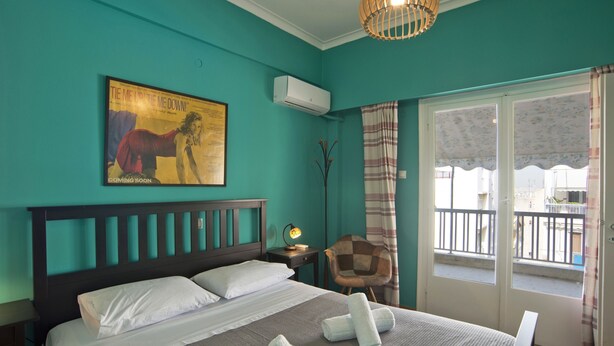 Une chambre aux murs de couleur turquoise, un lit avec du linge de lit blanc, un climatiseur fixé au mur, une affiche encadrée accrochée au mur, une chaise et un balcon avec vue.