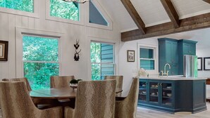 A dining area with a wooden table and patterned chairs, a kitchen with teal cabinets, and a view of greenery through large windows. - Luxury cabin w/ chef kitchen & two living rooms (Highlands)