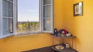 Habitación de esquina con un ventanal grande, una mesita con una taza, un hervidor y un florero con flores.