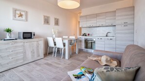 A modern kitchen with white cabinets, a dining area with white chairs, and a living area with a sofa and a magazine. - Sardinia Re - Smeralda 16 (Castelsardo)