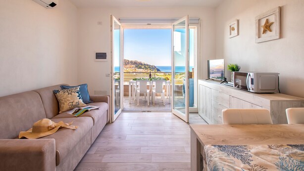 A modern living room with a sofa, a dining table, and a view of the sea through an open door. - Sardinia Re - Smeralda 16 (Castelsardo)