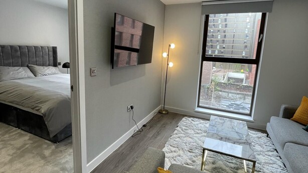 A modern living room with a bed, a flat-screen TV, a floor lamp, and a large window with a city view. - 5 Star Brand New Liverpool City Centre Apartment (Liverpool)