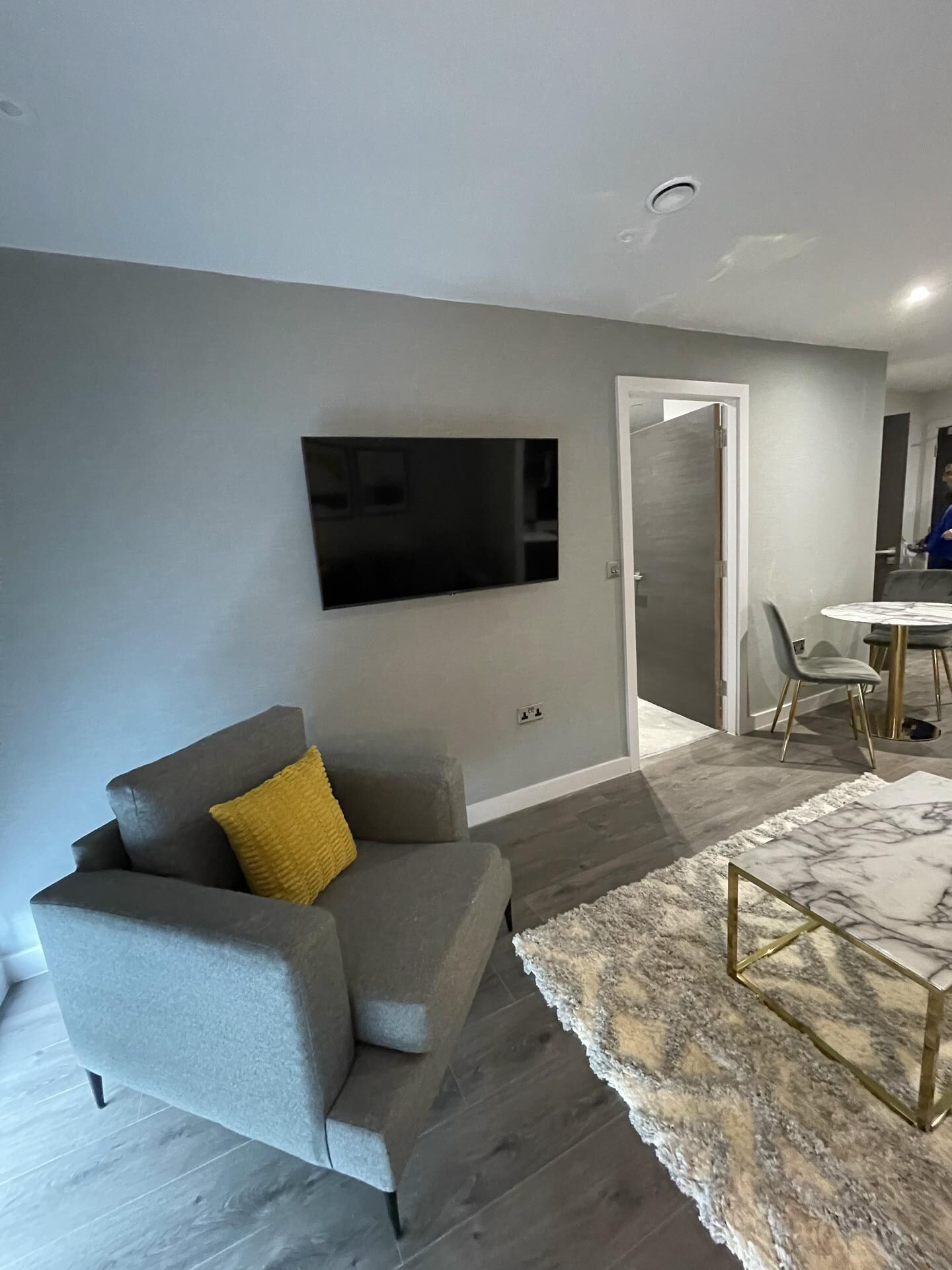 A modern living room with a grey armchair, a flat-screen TV mounted on the wall, a marble-patterned rug, and a dining area with a glass table and chairs.