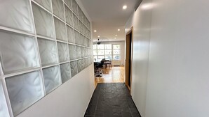 Een gang met een betegelde muur en een donkere vloer die naar een kamer leidt, met een tafel en stoelen.