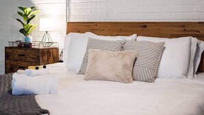 Una cama bien hecha con almohadas a rayas y lisas, un cabecero de madera y una mesita de noche con una lámpara y una planta.