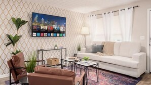 Una sala de estar con sofá blanco, una mesa de centro, un televisor montado en la pared que muestra varios servicios de streaming y un papel tapiz con diseño.