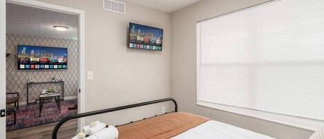 Habitación de hotel con cama, televisión montada en la pared, ventana con persianas y puerta que da a otra habitación.