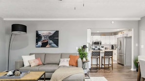 A modern living room with a grey sofa, a coffee table, and a painting of a lake. A kitchen is visible in the background with white cabinets and stainless steel appliances. - Suburban oasis perfect for families & groups (Columbus)