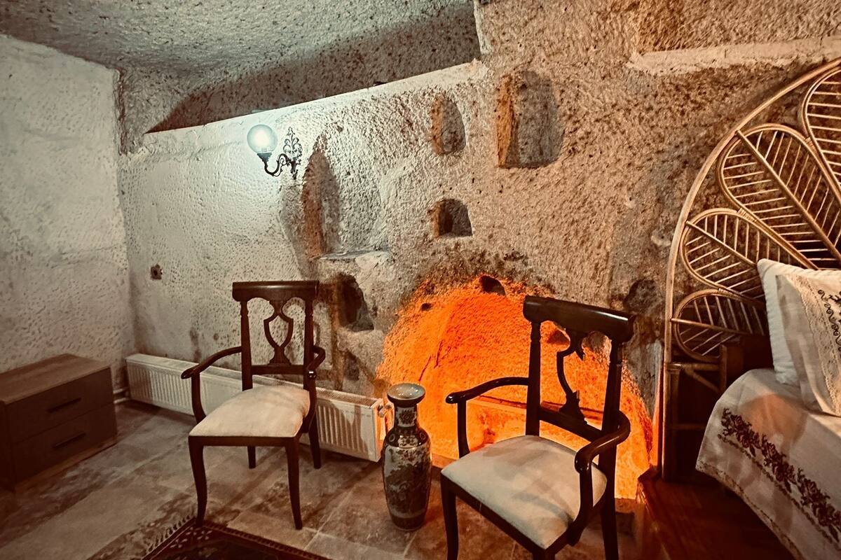 A cozy room with a stone wall, two wooden chairs, a small table, a decorative rug, and a bed with a patterned bedspread.