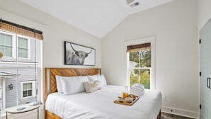 A bedroom with a wooden headboard, white bedding, a bedside table with a lamp, a framed picture on the wall, and a window with a view of greenery.