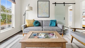 A living room with a sofa, a coffee table with magazines, a map on the wall, and a window with a view of the outside.