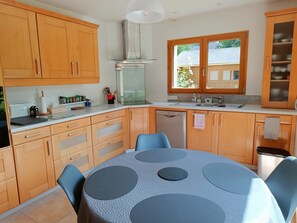 A kitchen with wooden cabinets, a dishwasher, a sink, and a dining table with chairs. - Quiet, bright single-storey house close to beaches, port and GR34. (Moëlan-sur-Mer)