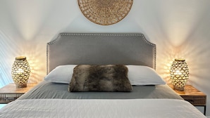 Ein Schlafzimmer mit grauem Polsterkopfteil, einer weißen gesteppten Bettdecke, einem braunen Fellüberwurf und zwei an der Wand befestigten Lampen mit geometrischen Mustern.