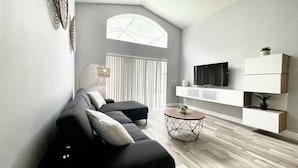 Ein modernes Wohnzimmer mit einer Couch, einem Couchtisch, einem an der Wand montierten Fernseher und einem großen Fenster mit vertikalen Jalousien.