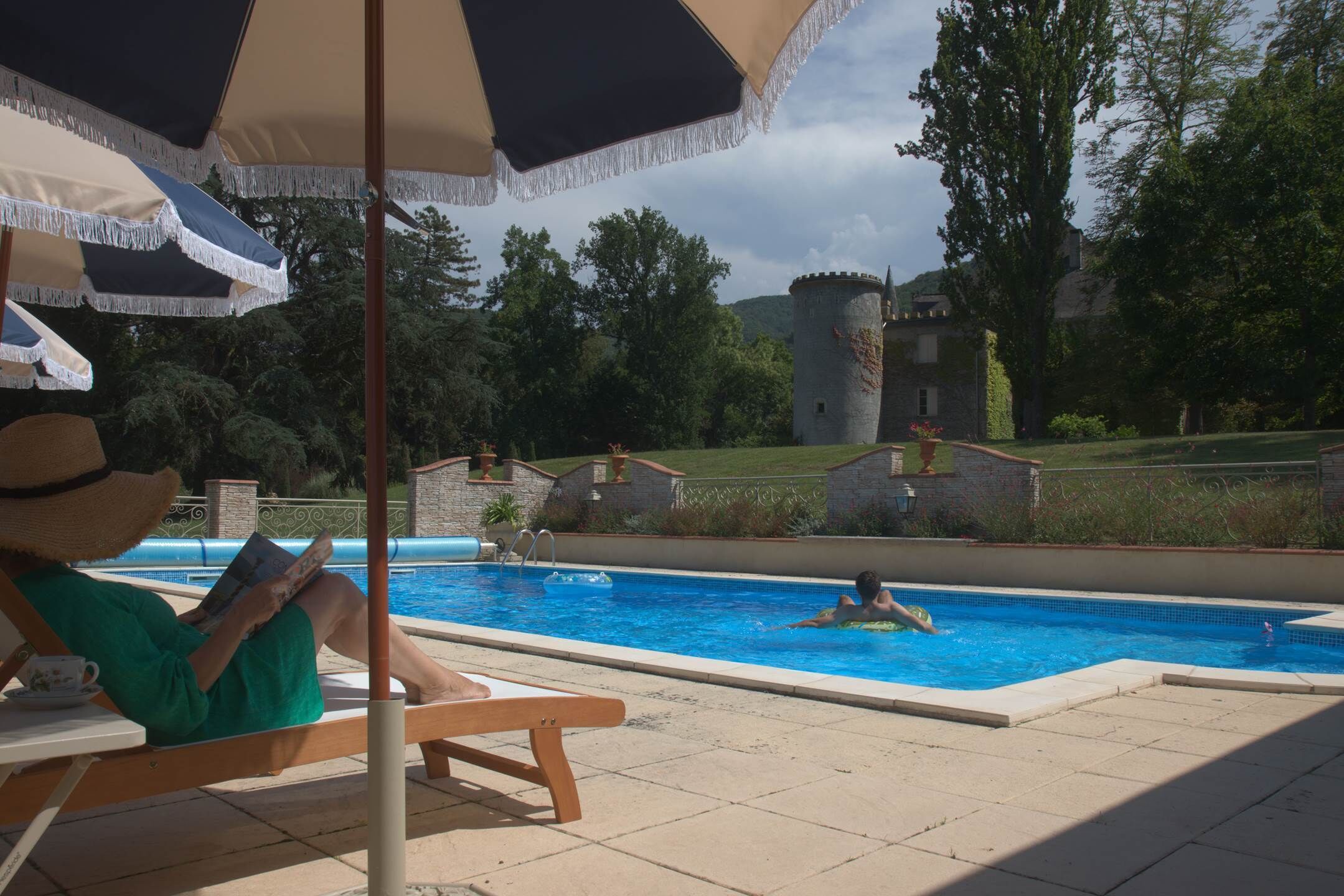 A pool area with a castle-like structure in the background, a person reading under an umbrella, and another person swimming.