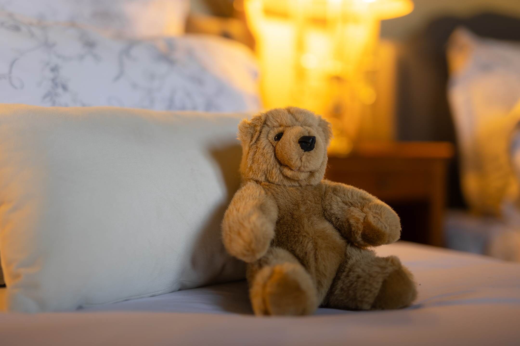 A teddy bear sitting on a bed with a white pillow and a lamp in the background.