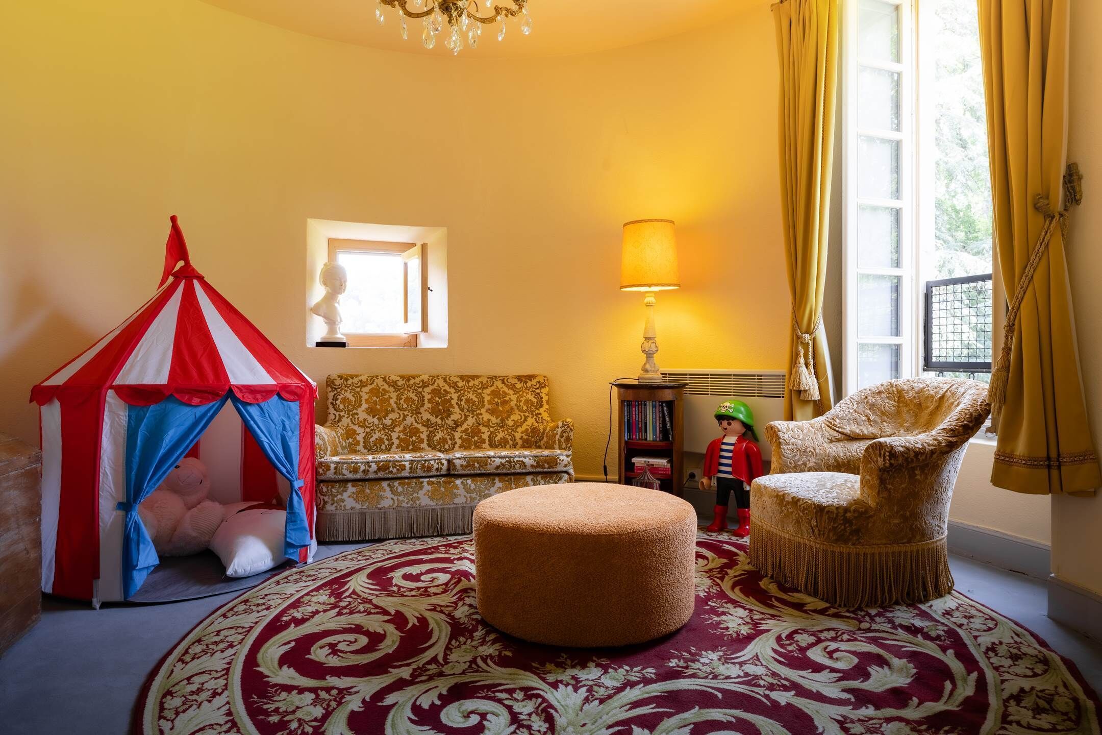A cozy room with a red and white striped tent, a round ottoman, a patterned sofa, a lamp, and a chair.