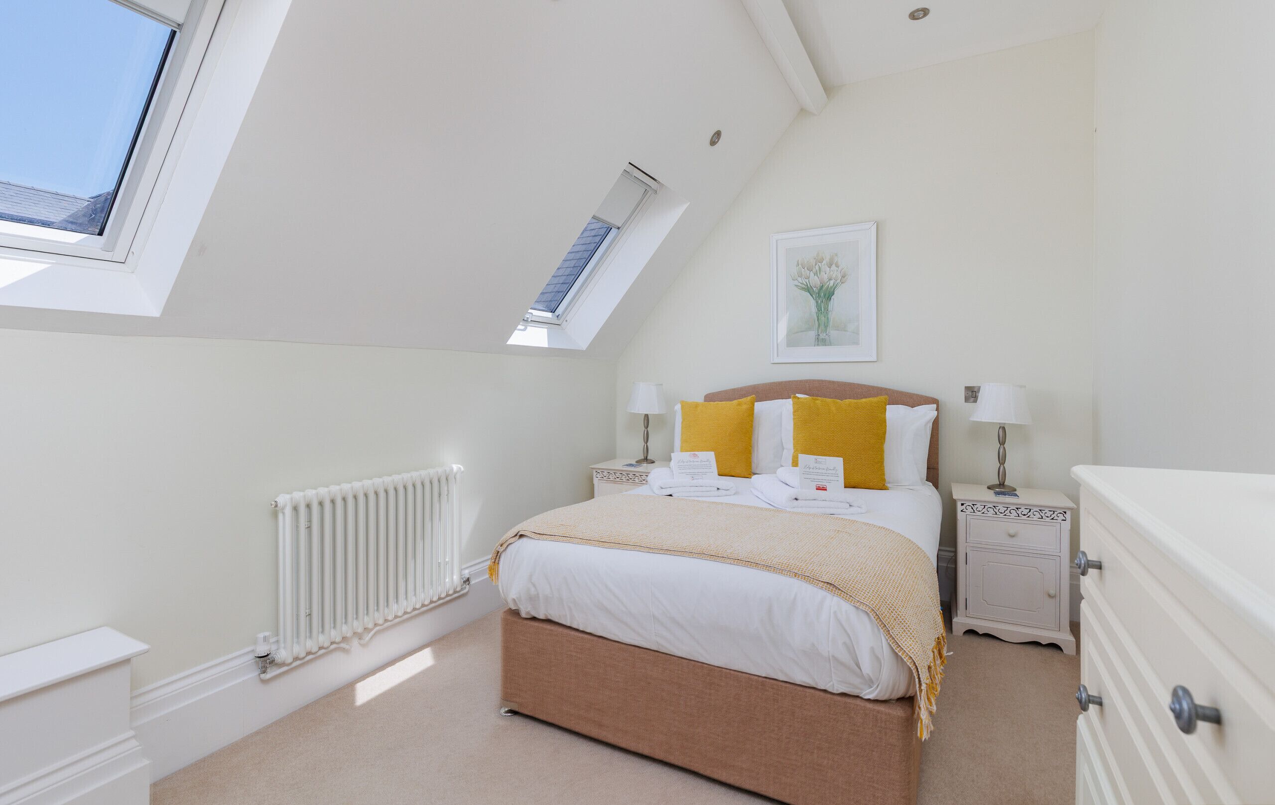 A bedroom with a skylight, a bed with yellow pillows, a bedside table with a lamp, and a dresser.