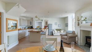 Una cocina moderna con armarios blancos, una zona de comedor con una mesa de madera, y una sala de estar con un sofá y sillones.