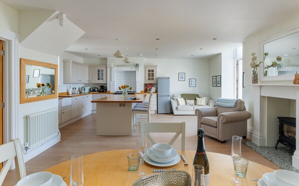 A modern kitchen with white cabinets, a dining area with a wooden table, and a living room with a sofa and armchairs.