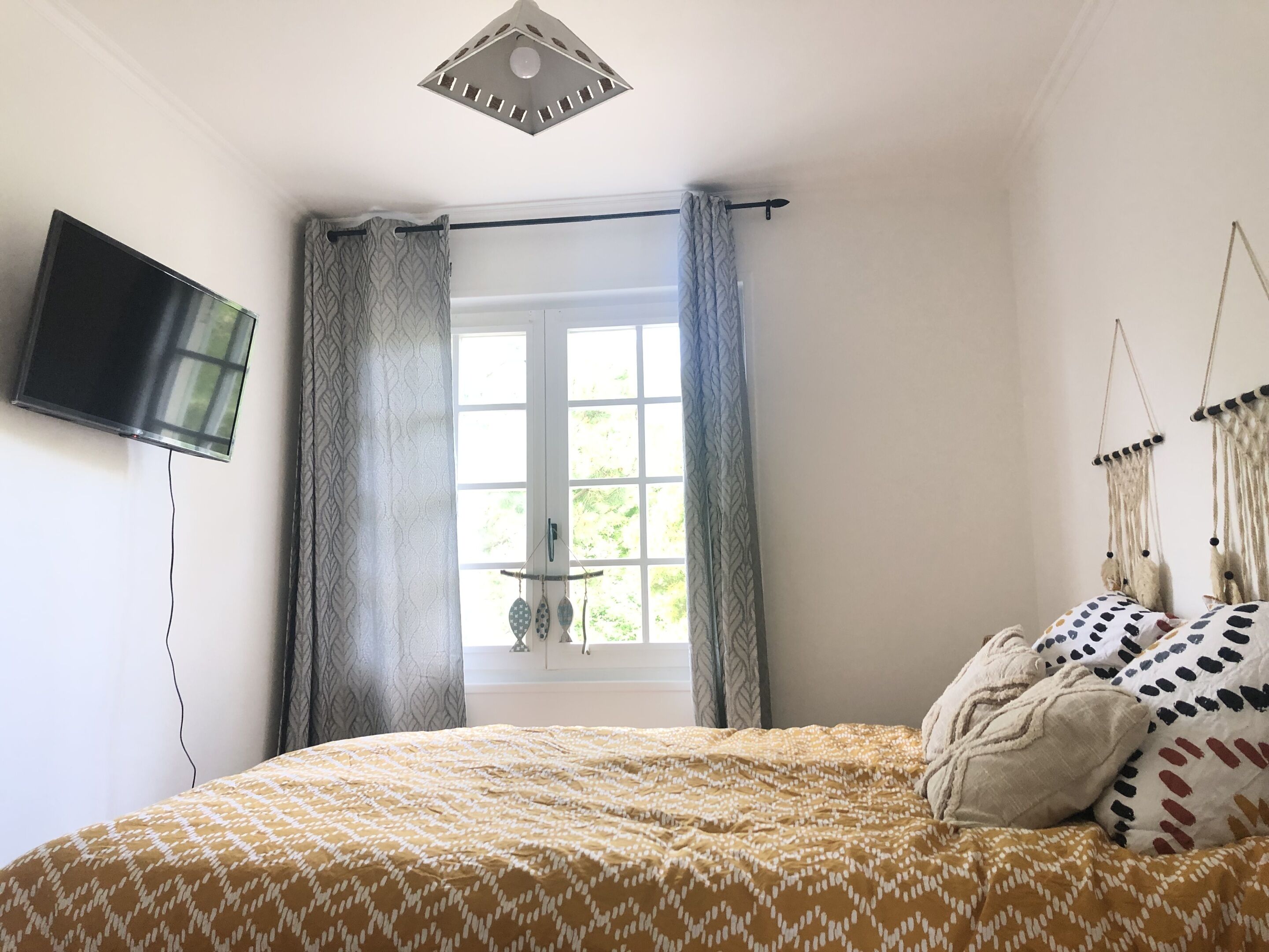 Une chambre à coucher avec un lit, un téléviseur fixé au mur, une fenêtre avec des rideaux et une décoration en macramé suspendue.