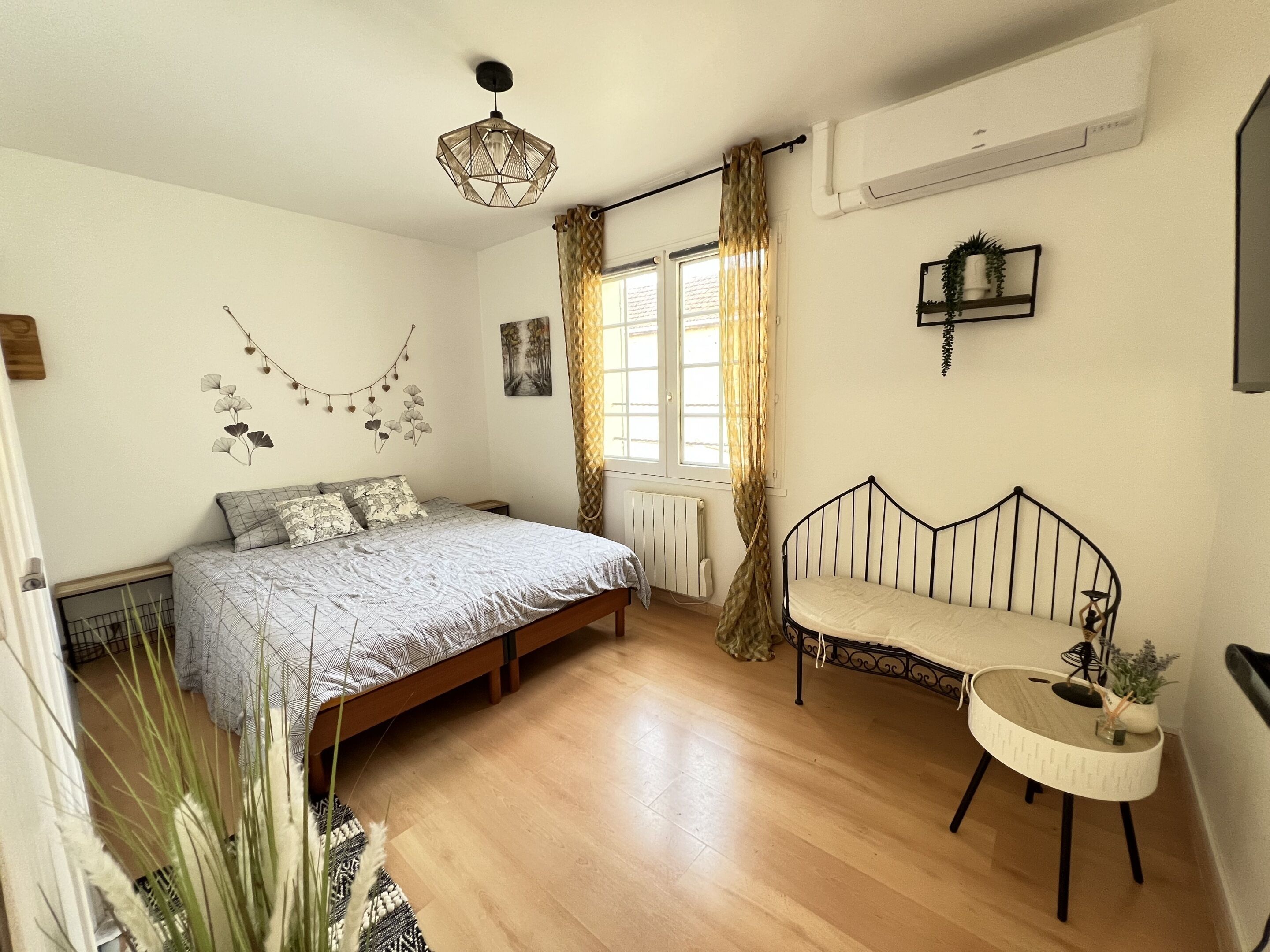A bedroom with a bed, a small bedside table, a chair, a window with curtains, a radiator, and a wall-mounted shelf.