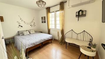 A bedroom with a bed, a small bedside table, a chair, a window with curtains, a radiator, and a wall-mounted shelf.