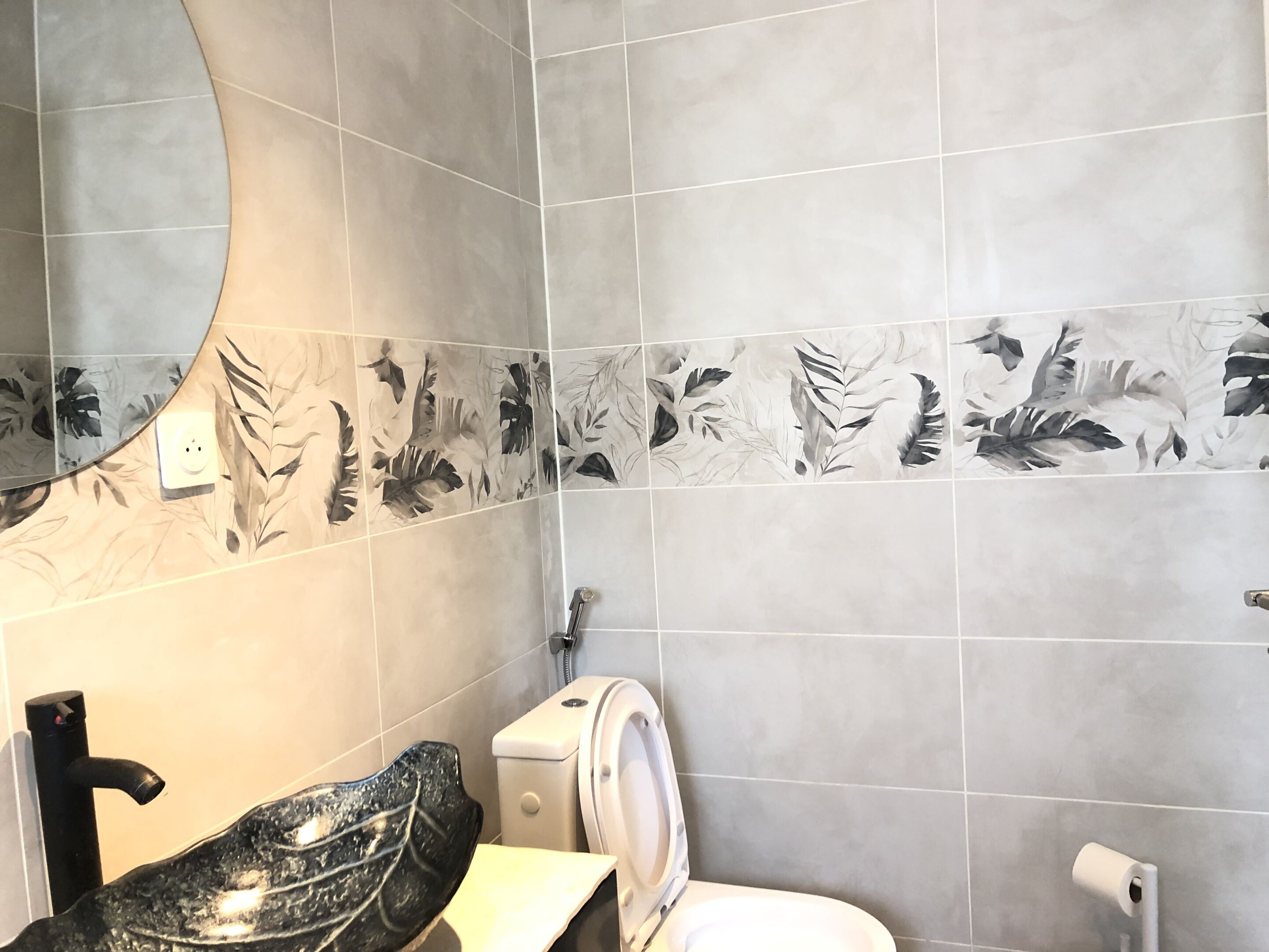 Et badeværelse med grå fliser, toilet, vask med sort armatur og en bruseniche.