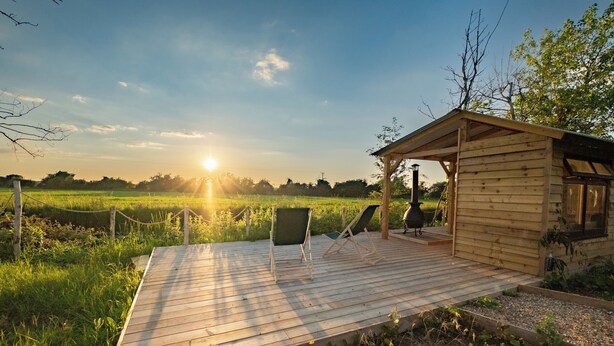 Une terrasse en bois dotée d’un espace salon couvert et d’un petit abri, donnant sur un champ verdoyant.