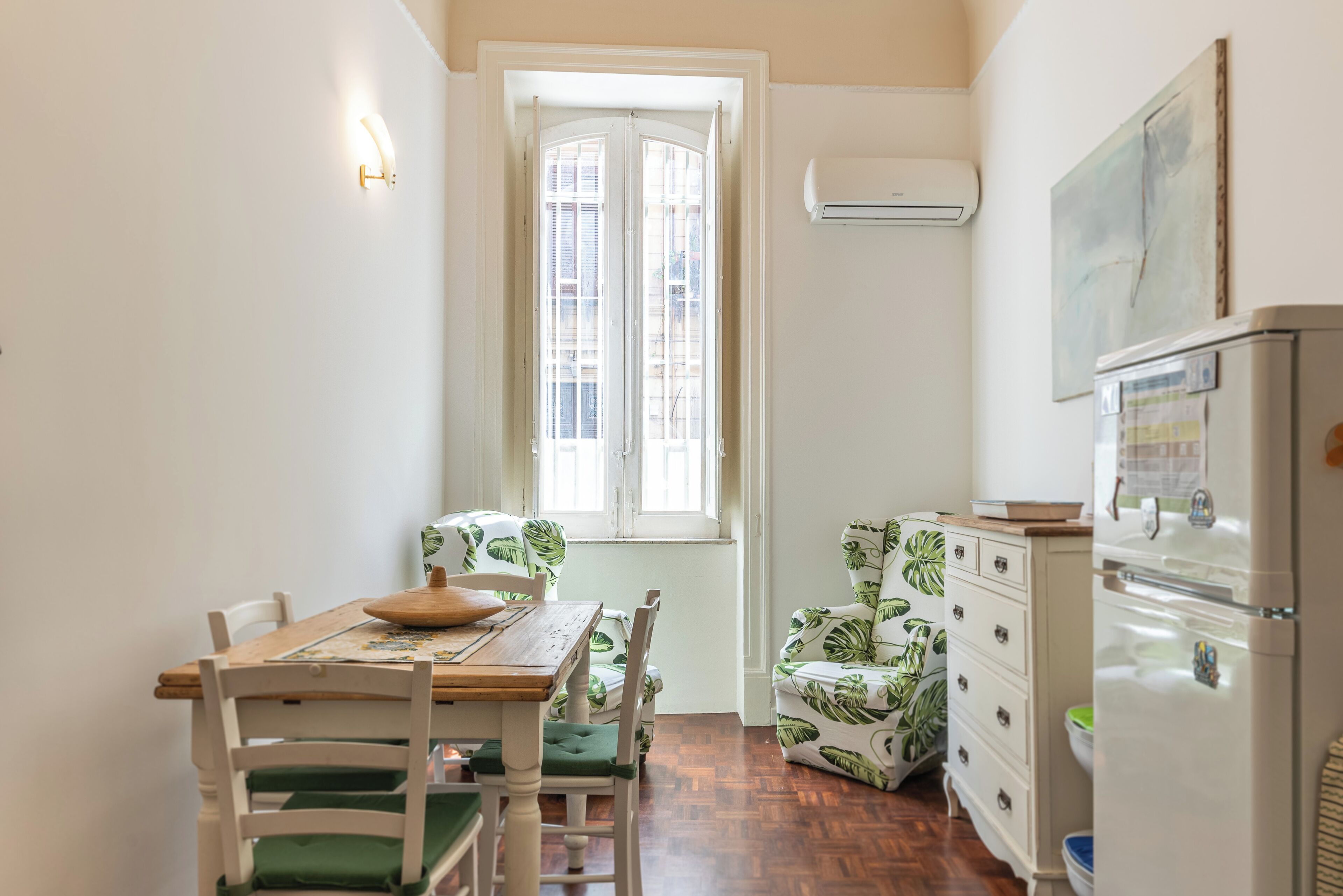 A compact kitchen with a dining table, chairs, a refrigerator, and a window with shutters.