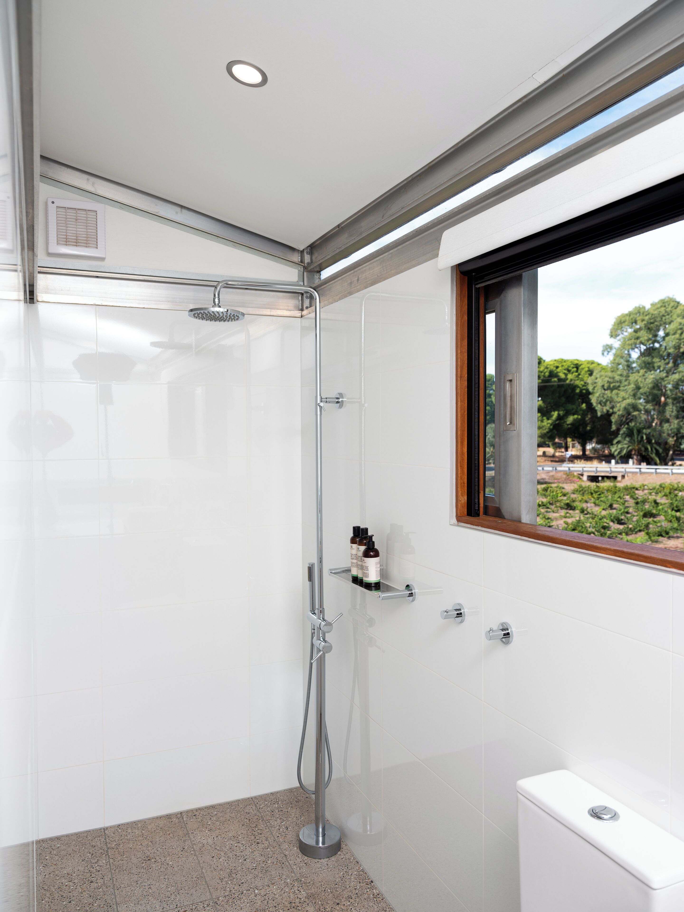 A modern bathroom with a glass-enclosed shower, white tiles, a toilet, and a window with a view of trees.