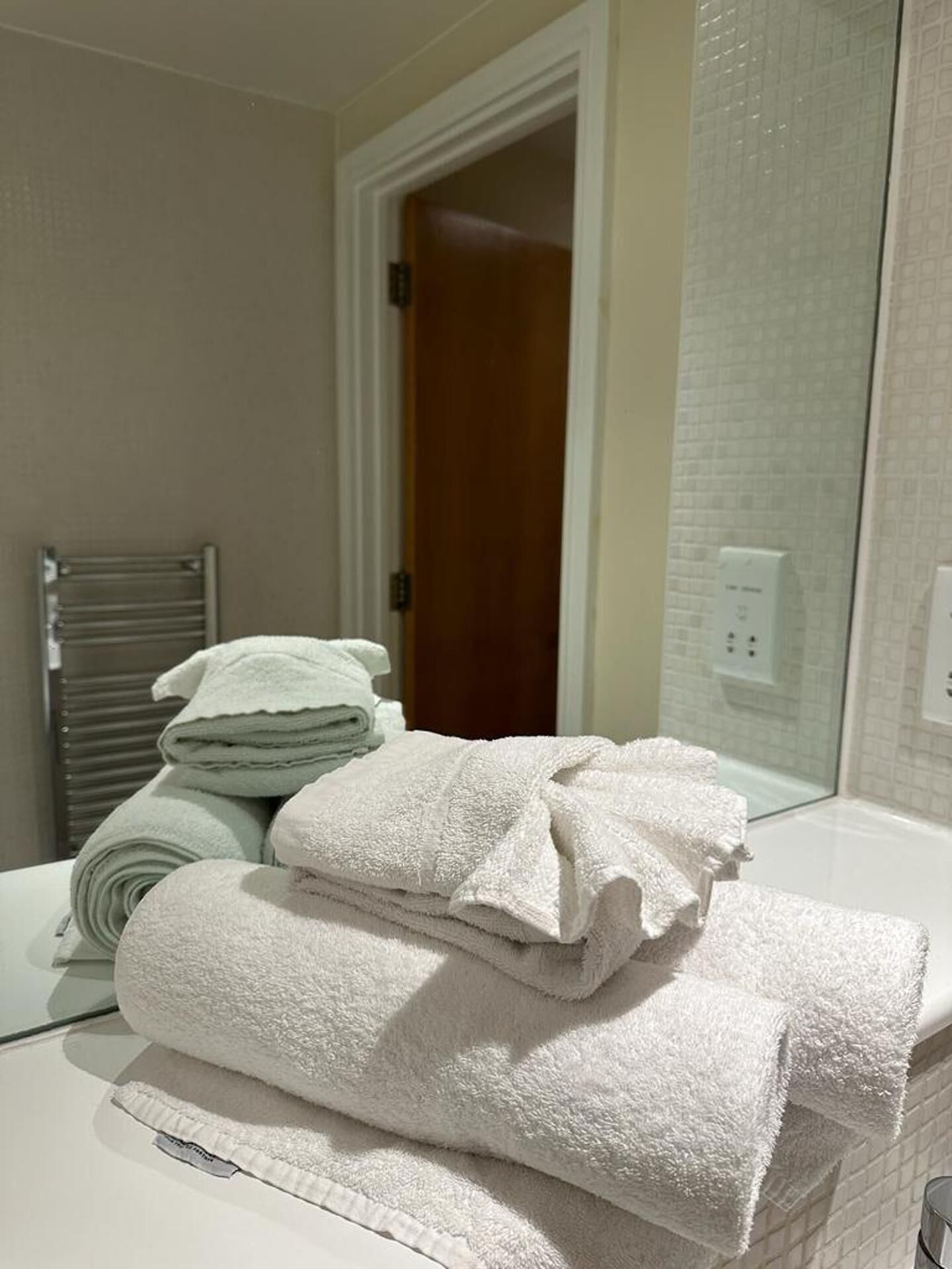A bathroom with a stack of white towels on a counter, a towel rack, and a wooden door in the background.