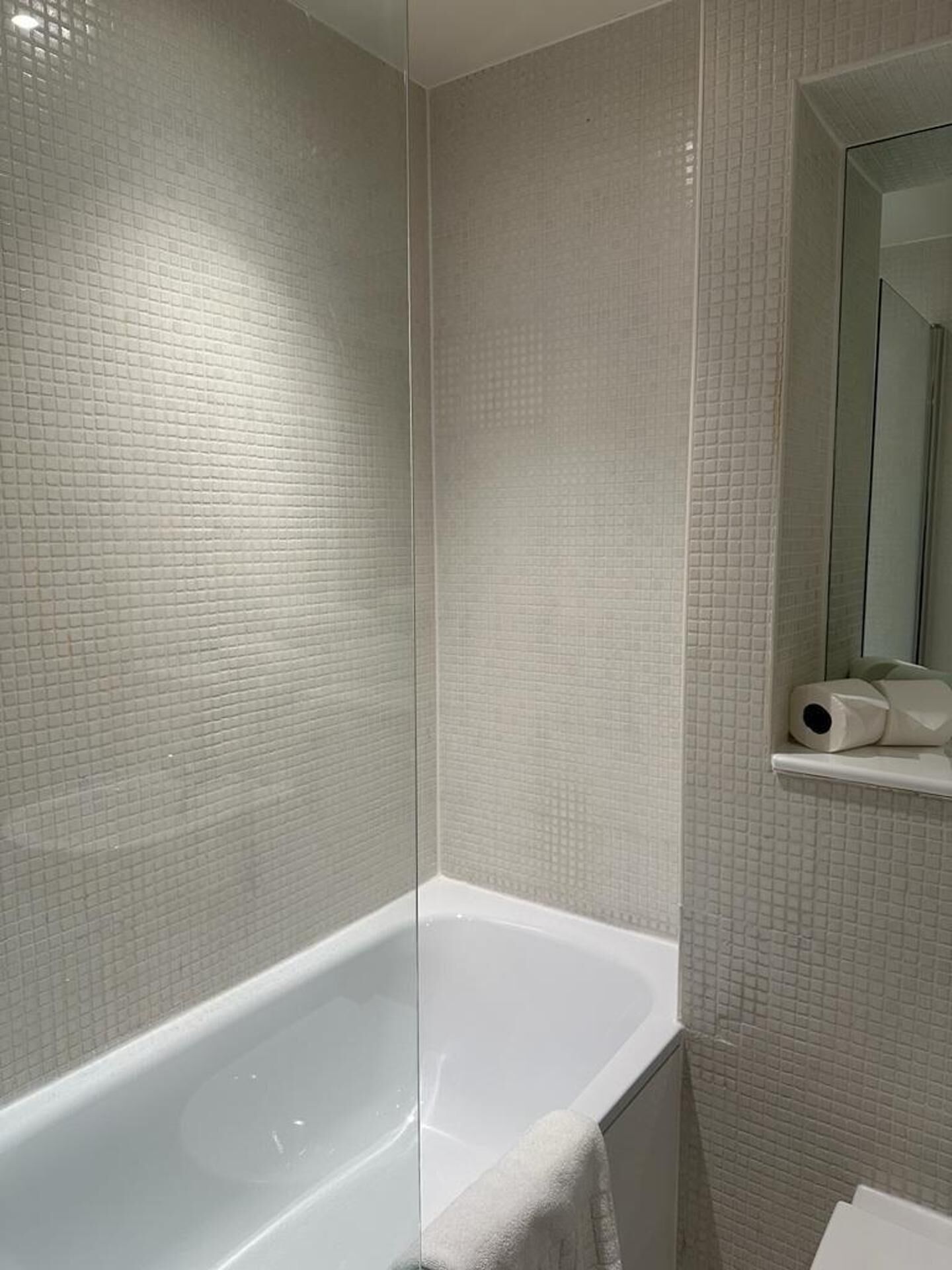 Un baño con una bañera blanca, paredes enlosadas y un espejo con un estante que sostiene un rollo de papel higiénico.