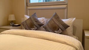 Habitación de hotel con cama, almohadas, mesita de noche, lámpara y ventana con vista a un edificio.