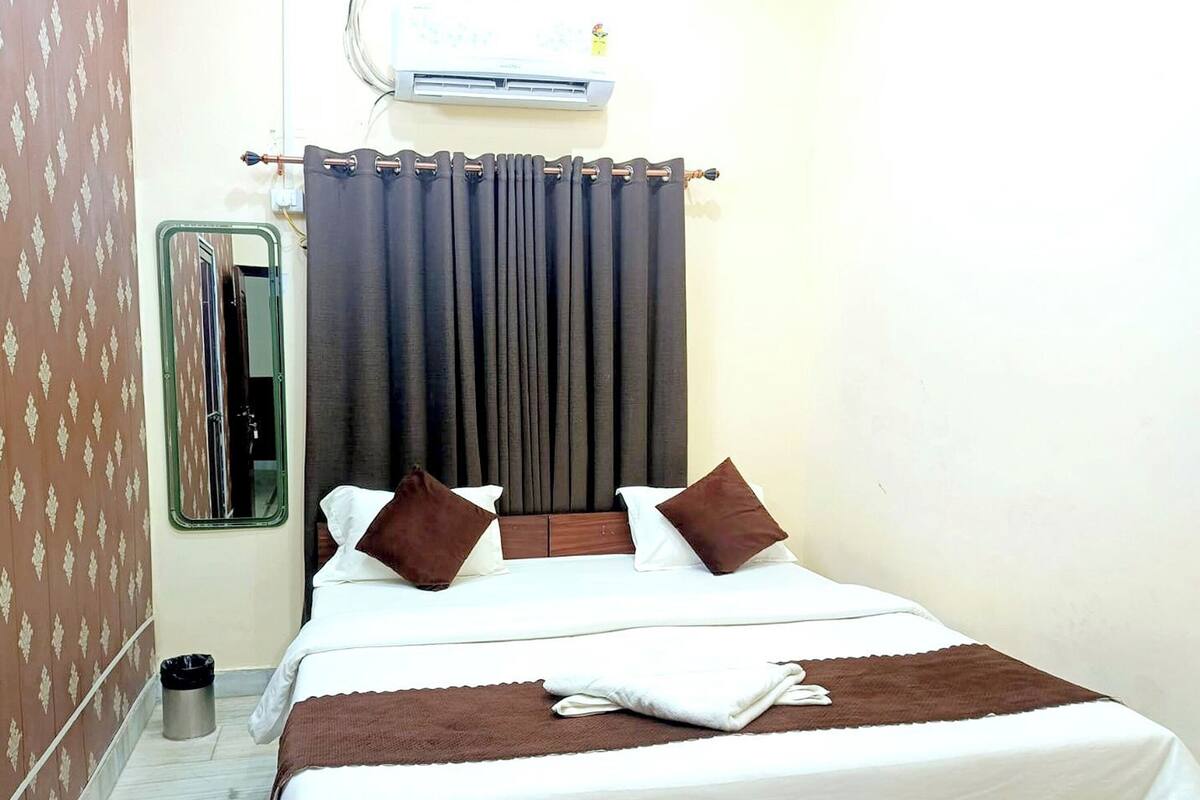 A hotel room with a bed, a window with a mirror, a wall-mounted air conditioner, and a trash bin.