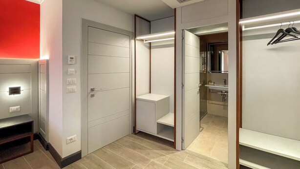 Ein modernes Hotelzimmer mit begehbarem Kleiderschrank, Schminktisch mit Spiegel und einer roten Akzentwand.